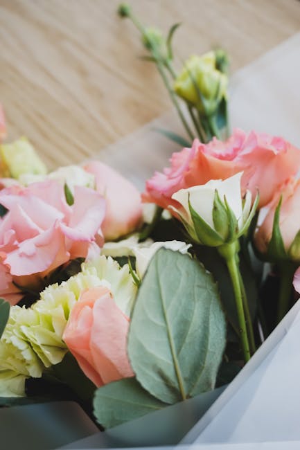 A close-up of a floral bouquet featuring soft pink and cream lisianthus flowers with delicate, ruffled petals, accented by fresh green leaves and buds. The flowers are wrapped in light, semi-transpare