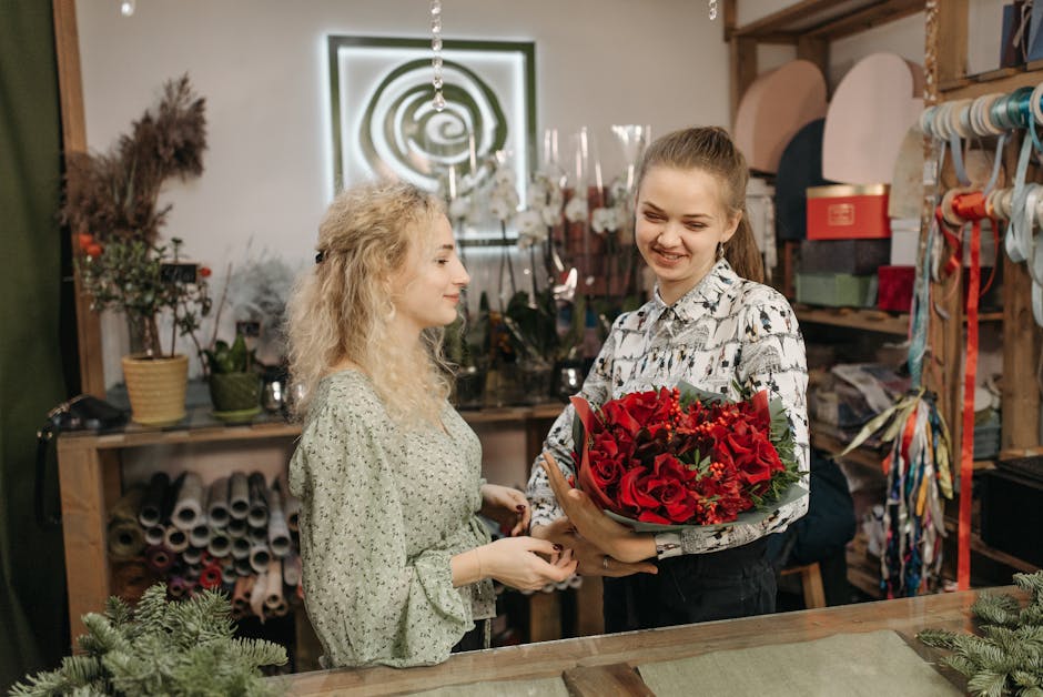 A young woman with light brown hair tied back, wearing a black and white patterned shirt, is handing a vibrant bouquet of red roses and accompanying red flowers, wrapped in dark green foliage and grey